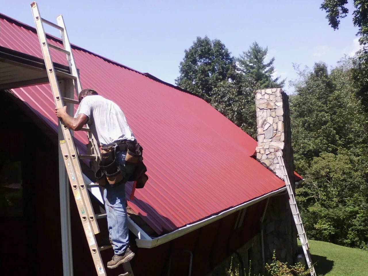 Roofer on a ladder beside a red metal roof and stone chimney — Soffit & Fascia Repair by Roofing Brothers in Cassoday
